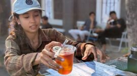 Aprodita saat menyajikan kopi untuk pelanggannya di atas sepeda roda tiga rakitan yang diubah menjadi meja barista, Rabu (22/7/26) pagi.